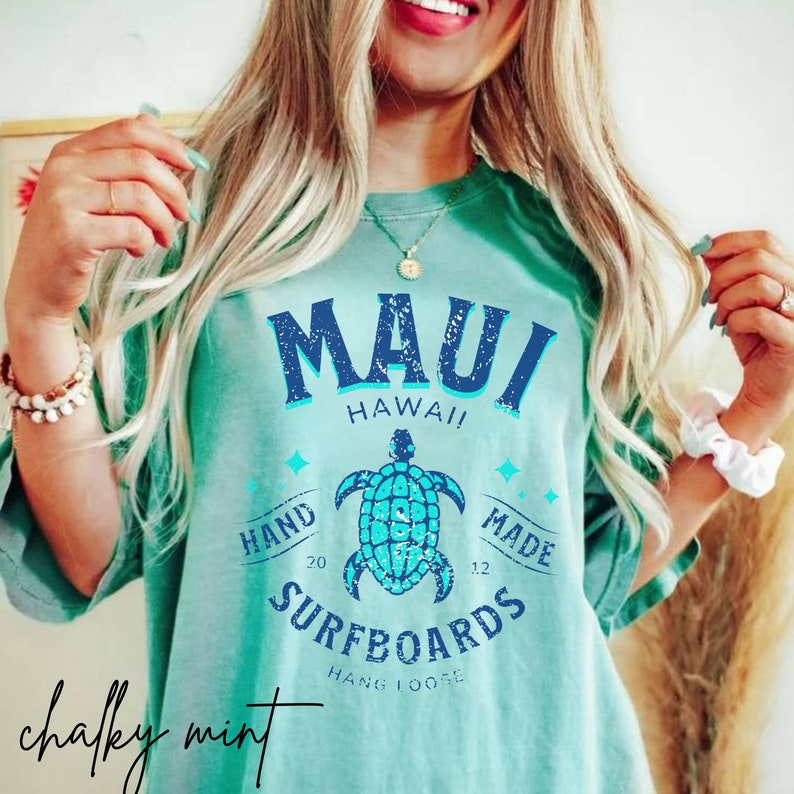 Pode incluir: Uma camiseta azul turquesa com um gr&aacute;fico azul desbotado que diz "Maui Hawaii" com uma tartaruga marinha e o texto "Hand Made Surfboards Hang Loose" em um estilo vintage.