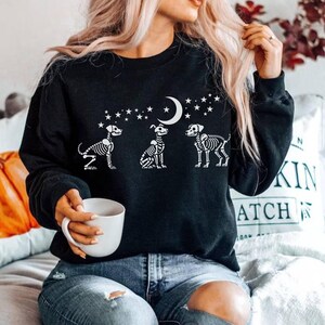 May include: Black sweatshirt with a white design of three skeleton dogs looking up at a crescent moon with stars. The sweatshirt is worn by a person with long blonde hair and blue jeans.