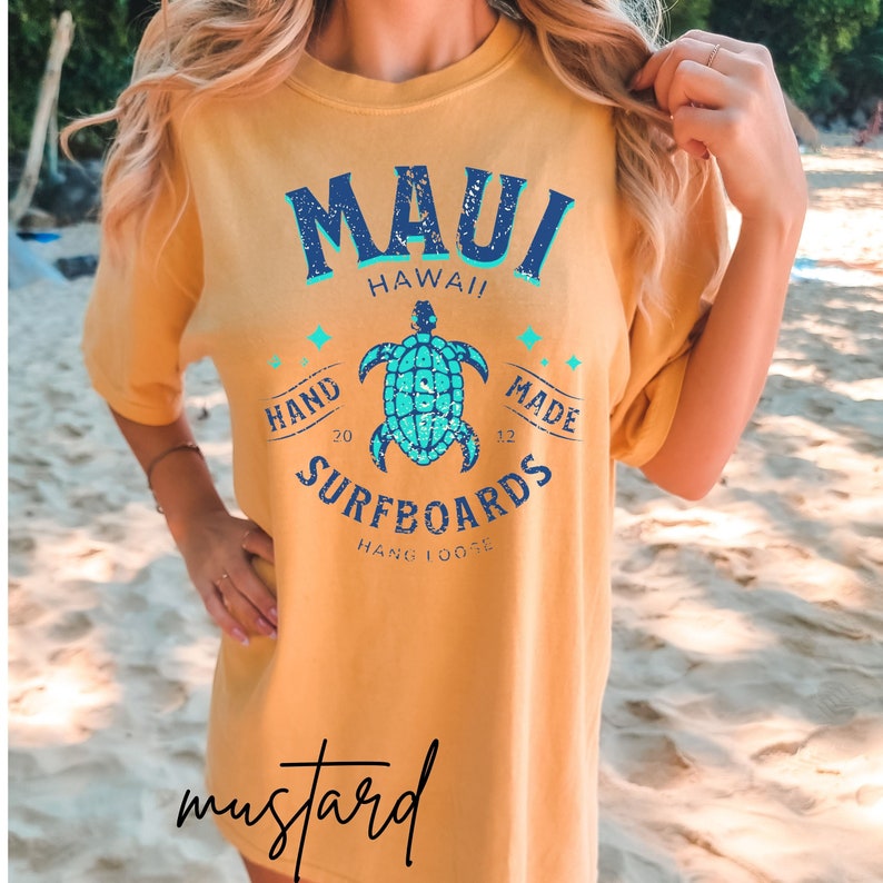 Pode incluir: Uma camiseta amarela mostarda com um design gr&aacute;fico que apresenta uma tartaruga marinha azul e turquesa, as palavras "Maui Hawaii", "Hand Made", "Surfboards" e "Hang Loose".