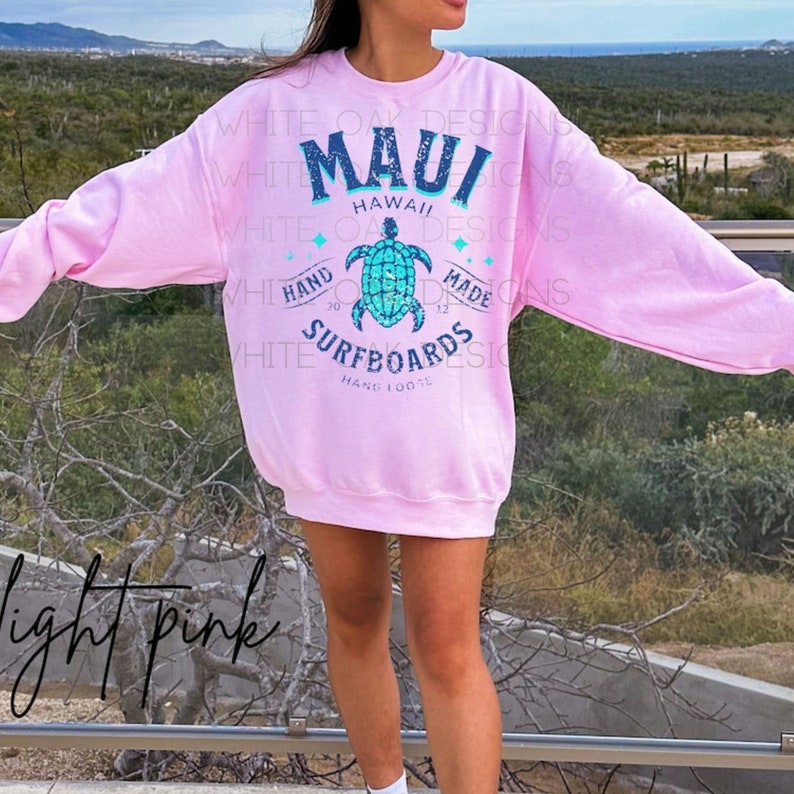 May include: A light pink sweatshirt with a graphic design featuring a sea turtle and the text "MAUI HAWAII HAND MADE SURFBOARDS HANG LOOSE".