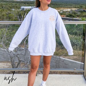 May include: A gray sweatshirt with a graphic of a sun setting over the ocean and the text "good vibes" in orange. The sweatshirt is being worn by a person standing in front of a railing.
