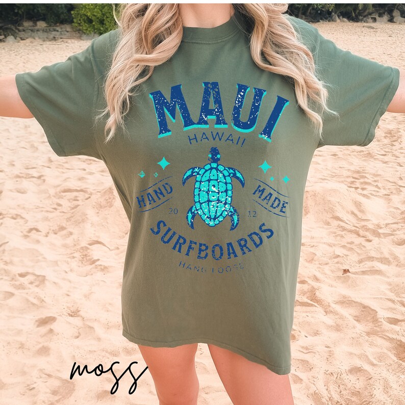 Pode incluir: Camiseta verde com um gr&aacute;fico azul e branco de uma tartaruga e o texto "MAUI HAWAII HAND MADE SURFBOARDS HANG 10"