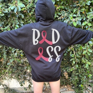 May include: Black hooded sweatshirt with the text "BADASS" in white with pink ribbon accents.