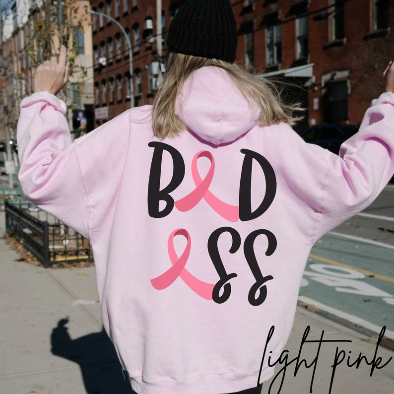 May include: Pink hoodie with a black graphic that says "BADASS" with pink ribbon accents. The hoodie is light pink.