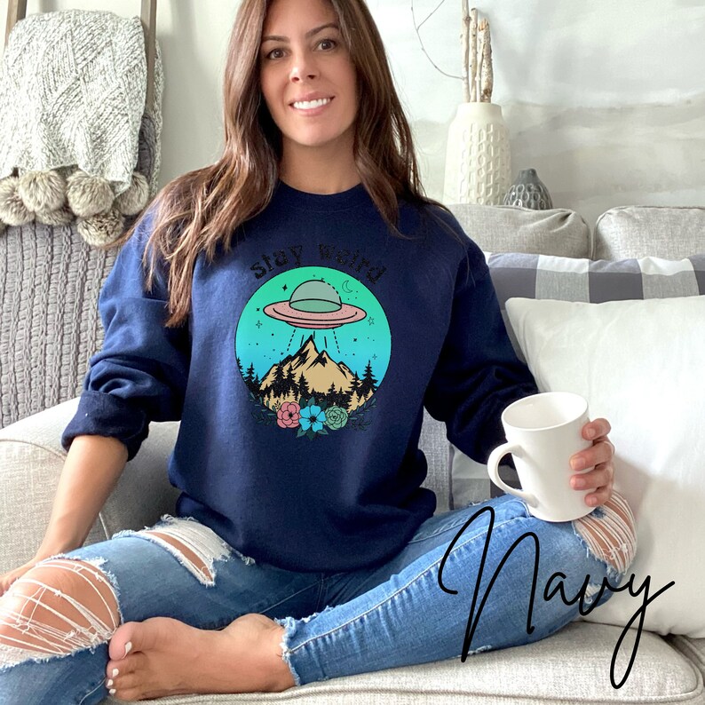 May include: A navy blue sweatshirt with a graphic of a UFO flying over a mountain range with a forest and flowers. The text "Stay Weird" is above the graphic.