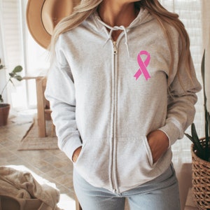 May include: A gray zip-up hoodie with a pink ribbon on the left chest. The hoodie has a hood and two pockets.