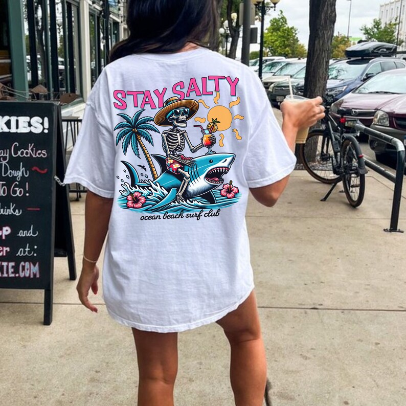 May include: White t-shirt with a graphic of a skeleton riding a shark with the text "STAY SALTY" and "ocean beach surf club".