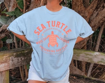 Puede incluir: Camiseta azul claro con un gráfico de una tortuga marina y el texto "Sea Turtle Conservation, Wave of Change, Preserving Our Seas, From Coral Reefs to Sandy Shores".