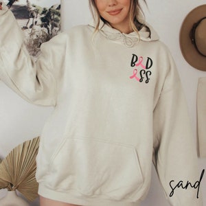 May include: A light beige hoodie with a black graphic that says "BADASS" with two pink ribbons.