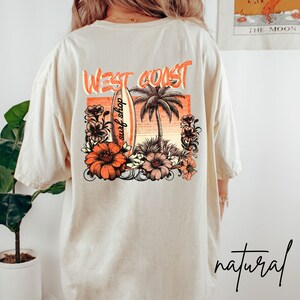 May include: A beige t-shirt with a graphic print of a surfboard, palm trees, and flowers. The text "West Coast surf shop" is printed on the surfboard. The text "natural" is printed in black at the bottom of the image.