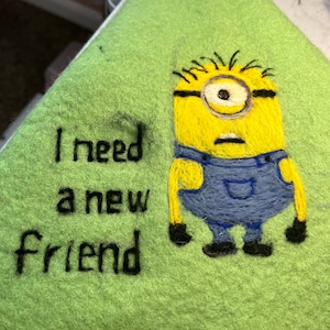 May include: A green felt item with embroidered text reading "I need a new friend." A yellow Minion character in blue overalls is embroidered on the right side of the felt.
