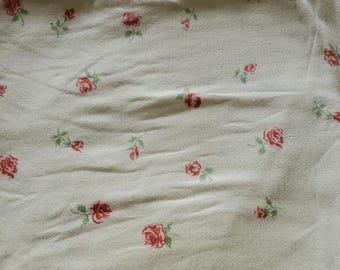 May include: Cream-colored fabric with a repeating pattern of small red roses and green leaves. The roses are scattered across the surface, creating a delicate floral design. The fabric appears soft and slightly textured.