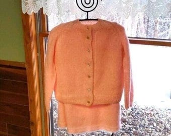 May include: A vintage peach-colored knit cardigan and skirt set. The cardigan has a round neckline and gold-colored buttons. The skirt is a matching peach color. The set is displayed on a black metal hanger in front of a window.