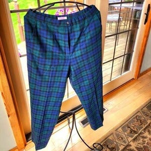 May include: Blue and green plaid pants hanging on a black hanger. The pants have a button closure and pockets. The plaid pattern is a mix of blue, green, and black. The pants are made of a woven fabric.