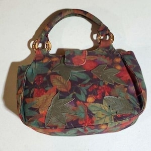 May include: A patterned handbag with a fall leaf design. The bag has a handle, a flap closure, and leaf-shaped appliqués. The colours include red, green, and brown, evoking an autumnal theme.