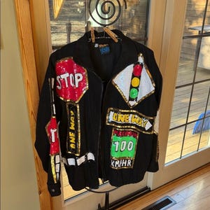 May include: Black jacket with colorful sequined road sign designs, including a red "STOP" sign, a traffic light, and a speed limit sign. The jacket has a zipper closure and the text "ONE WAY".