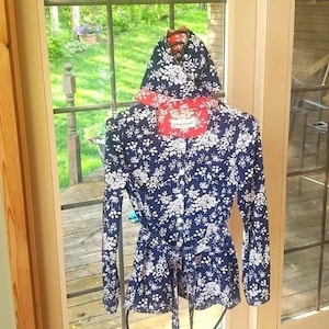 May include: A navy blue jacket with a white floral pattern, a hood, and a matching belt. The jacket has long sleeves and a red collar. The jacket is hanging on a wooden hanger in front of a window.