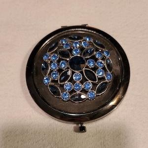 May include: A round, black compact mirror with a decorative design of blue and dark blue rhinestones. The rhinestones are arranged in a floral pattern. The compact is closed and has a small hinge and clasp.