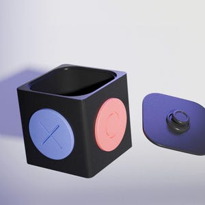 Könnte beinhalten: Ein schwarzer, quadratischer Behälter mit passendem Deckel. Der Behälter hat ein blaues X und ein rosa O an den Seiten. Der Deckel ist dunkelviolett und hat einen kleinen Knopf.