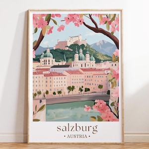May include: A framed art print depicting Salzburg, Austria, with a scenic illustration. The artwork shows a cityscape with buildings, a river, and a mountain backdrop. Pink blossoms frame the view, with the city's name and country at the bottom.