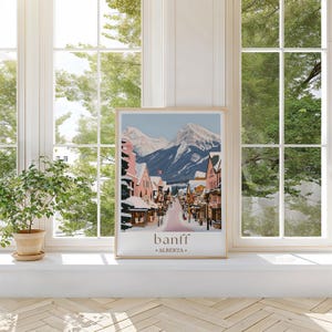 A framed poster of a snowy mountain town in Banff, Alberta, Canada, displayed on a white shelf in front of a window with trees visible outside.