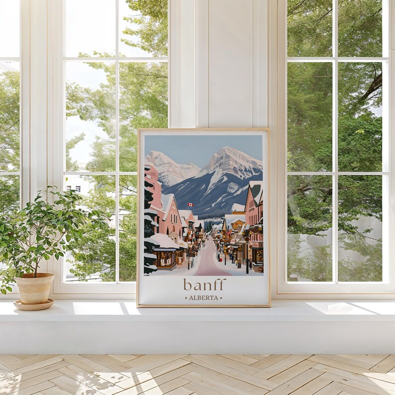 A framed poster of a snowy mountain town in Banff, Alberta, Canada, displayed on a white shelf in front of a window with trees visible outside.