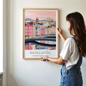 A woman is hanging a framed poster of Newcastle, United Kingdom, on a wall.