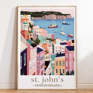 Op de afbeelding: Een ingelijste kunstdruk met een kleurrijke illustratie van St. John's, Newfoundland. Het kunstwerk toont een kustplaats met levendige gebouwen in roze, blauwe en gele tinten, met boten op het water. De tekst "st. john's • NEWFOUNDLAND" staat onderaan.