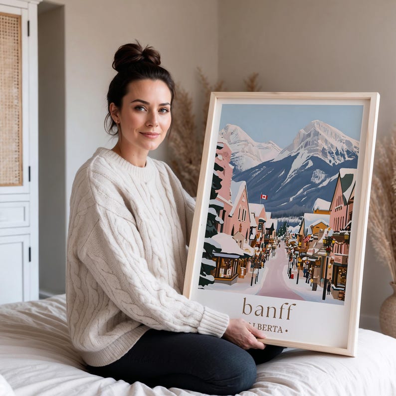 A woman in a white sweater is sitting on a bed and holding a framed poster of a snowy mountain town.