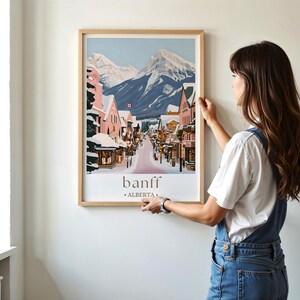 A woman is hanging a framed poster of a snowy mountain town on a wall.