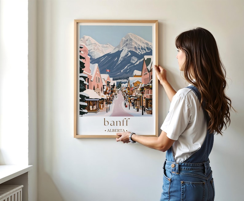 A woman is hanging a framed poster of a snowy mountain town on a wall.