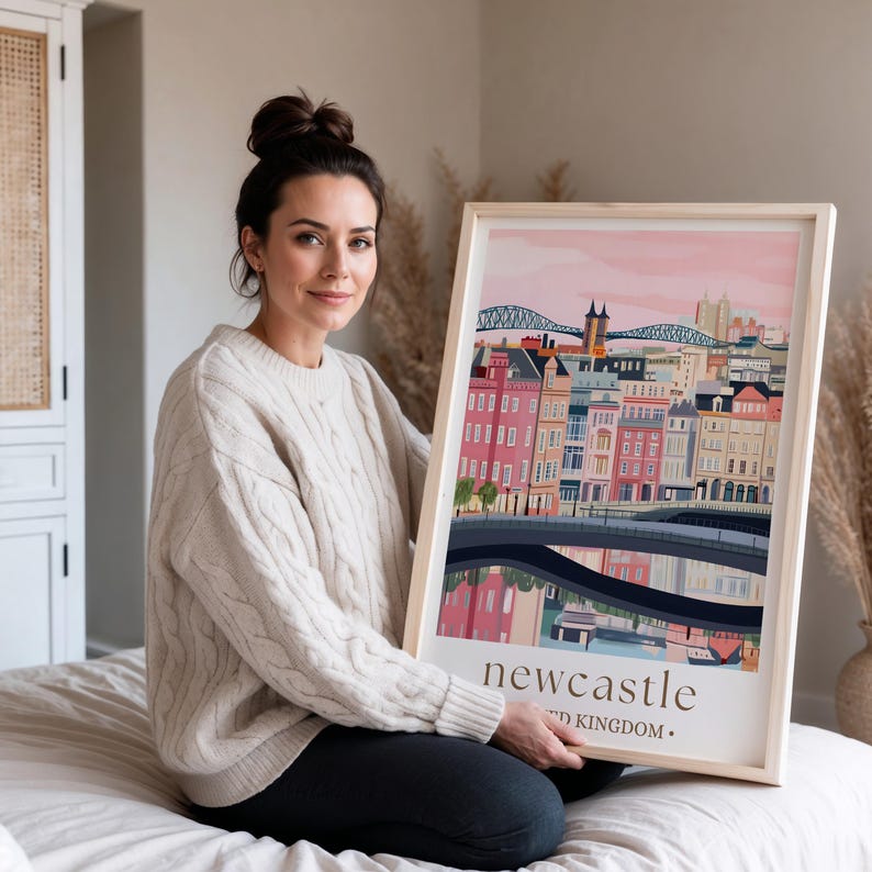 A woman is sitting on a bed and holding a framed poster of Newcastle, UK, with a cityscape illustration on it.