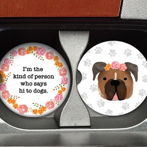 May include: Two round car coasters with a white background. One coaster features a floral wreath with the text "I'm the kind of person who says hi to dogs." The other coaster features a brown and white dog with a floral crown and paw prints.