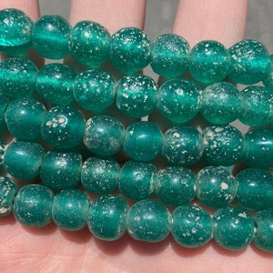 May include: A strand of green glass beads with a speckled texture. The beads are round and have a slightly irregular shape.