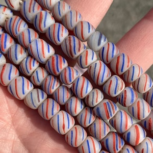 Puede incluir: Una hebra de cuentas de vidrio blanco con rayas rojas y azules. Las cuentas son de forma ovalada y tienen un acabado brillante.