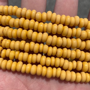 Op de afbeelding: Close-up van verschillende strengen gele kralen. De kralen zijn klein, cilindrisch en hebben een matte afwerking. De kleur is een zacht, gedempt geel. De kralen zijn aan elkaar geregen, waardoor een ketting of armband ontstaat.