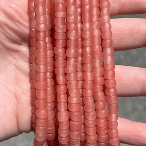 May include: Multiple strands of pink beads. The beads are small, cylindrical, and have a matte finish. The beads are strung together, forming multiple strands. The colour is a soft, muted pink, and the beads appear to be made of glass or a similar material.