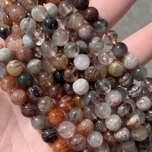May include: A strand of gemstone beads in various colors, including clear, brown, black, and orange. The beads are round and appear to be made of a translucent material, with visible inclusions. The beads are held in a hand, showcasing their natural beauty.