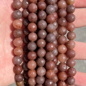 May include: A collection of faceted, round beads in varying shades of dusty rose, mauve, and earthy brown. Each bead features a multi-faceted surface that catches the light, creating a textured appearance across the multiple strands of gemstone beads.