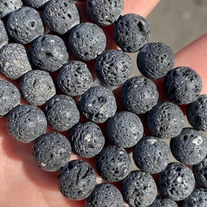 May include: Close-up of a collection of dark gray lava rock beads. The spherical beads have a porous texture and are arranged closely together. These beads are often used in jewelry making and aromatherapy.