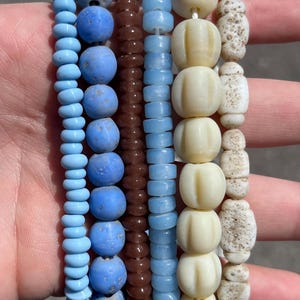 May include: A collection of beaded bracelets in various colors and styles. The bracelets feature beads in shades of light blue, brown, and cream. Some beads are round, while others have a ribbed or textured design. The bracelets are strung together on a white string.