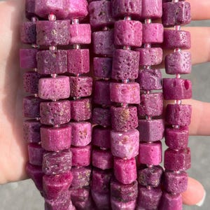 May include: A strand of raw, deep pink ruby beads shaped into irregular cubes. The beads feature a porous, textured surface and are strung together with small, clear spacer beads between each stone, creating a continuous, vibrant jewelry component.