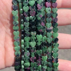 May include: Multiple strands of heart-shaped beads made from zoisite with ruby inclusions. The beads feature a mottled pattern of forest green, black, and vibrant magenta-pink, arranged in vertical rows to form a collection of gemstone jewellery components.