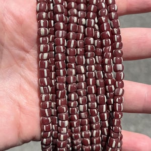 May include: A collection of maroon beads with white stripes. The beads are cylindrical and strung together, creating a textured pattern. The beads are held in a hand, showing the colour and design. Each bead is approximately 0.5 cm in length.