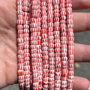 Può includere: Fili di perline a righe rosse e bianche, simili a caramelle alla menta piperita. Le perline sono cilindriche e infilate insieme, creando un motivo festoso e colorato. Le perline sono tenute in una mano.