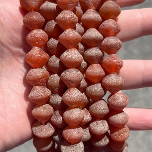 Peut inclure: Un fil de perles de pierres précieuses brun rougeâtre. Chaque perle a une forme unique, avec un centre arrondi et des bords facettés. Les perles ont un aspect légèrement translucide et une surface texturée. Les perles sont enfilées ensemble, créant un collier ou un bracelet.