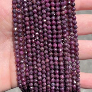 May include: A collection of multiple strands of small, round, faceted ruby beads. The beads are a deep, saturated magenta-purple colour with a reflective, multi-faceted surface texture. The strands are arranged in a parallel, vertical formation.