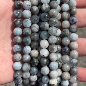 May include: A collection of smooth, spherical beads arranged in multiple vertical strands. The beads feature a marbled appearance with varying shades of gray, blue, and white, displaying natural inclusions and subtle variations in color and texture.