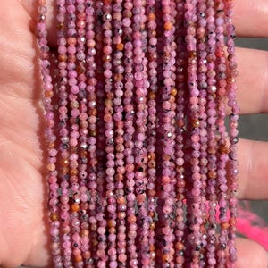 May include: Multiple strands of small, faceted, round pink sapphire beads. The beads display a range of pink and magenta hues with natural dark inclusions and reflective surfaces, arranged in parallel vertical lines to form a long, continuous display.