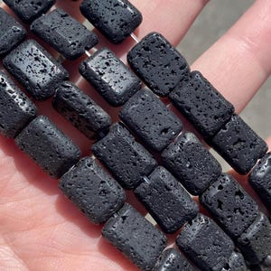 Puede incluir: Una hebra de cuentas de roca volcánica negra, cada cuenta es rectangular y tiene una textura rugosa y porosa.